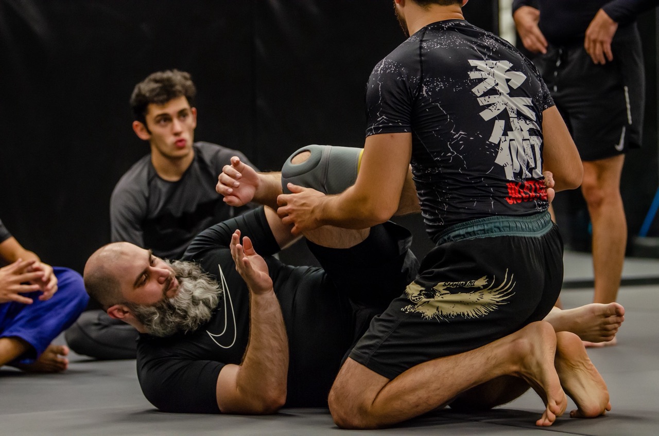 Two adult grapplers working a guard position on the mats while other athletes watch from the edge